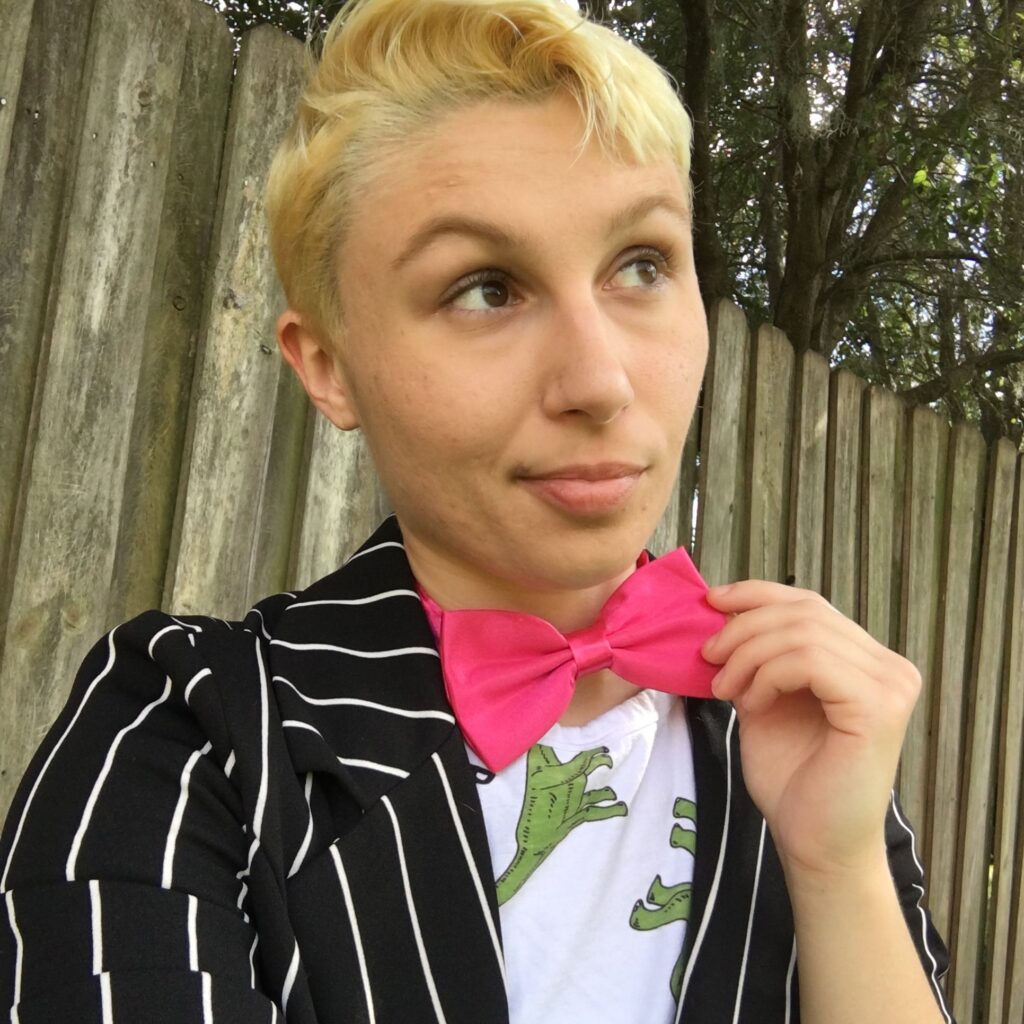 [IMAGE ID: A thin white masculine nonbinary person with short blonde hair & brown eyes looks off to the side of the camera. They are sitting outdoors in front of a wooden fence & wearing a white shirt with green dinosaurs on it, a black & white striped blazer, & a pink bow tie, which they are holding the edge of in their left hand. END ID]