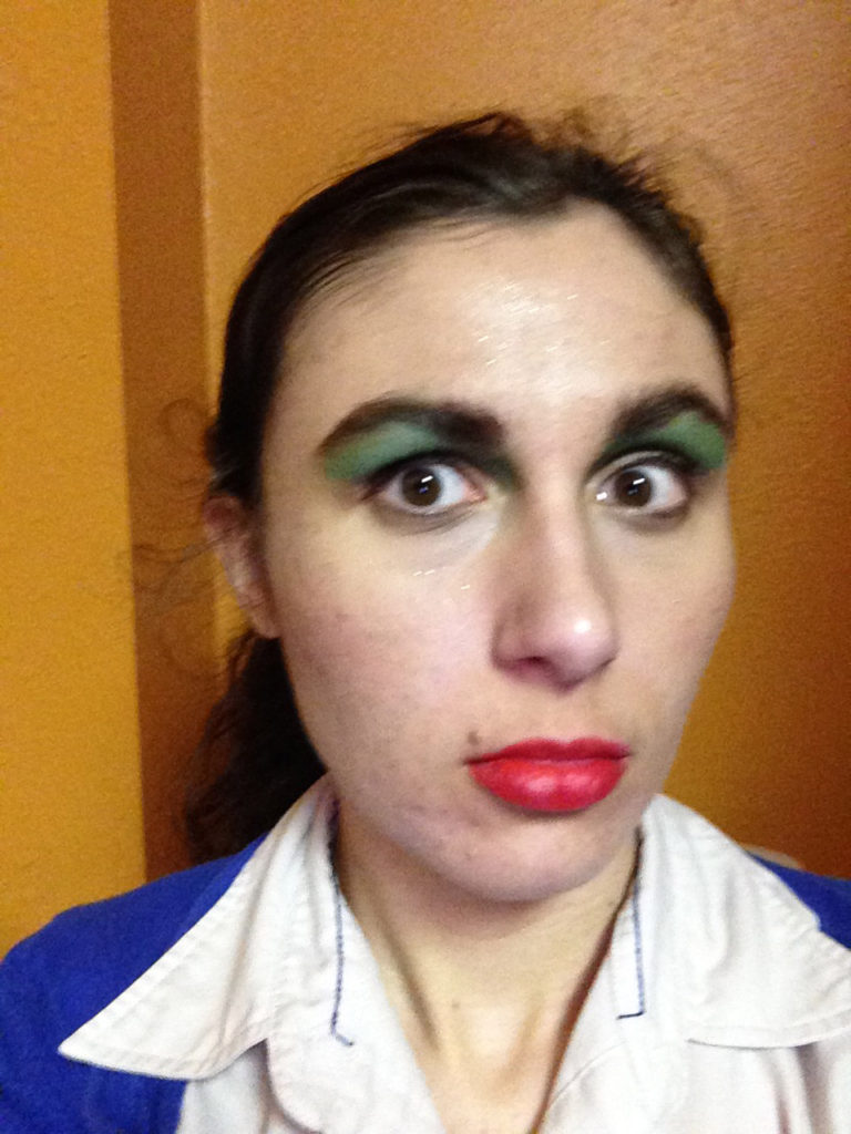 Ashlee Craft wearing a blue sweater, green eyeshadow, theatre makeup, a dress shirt, & ponytail